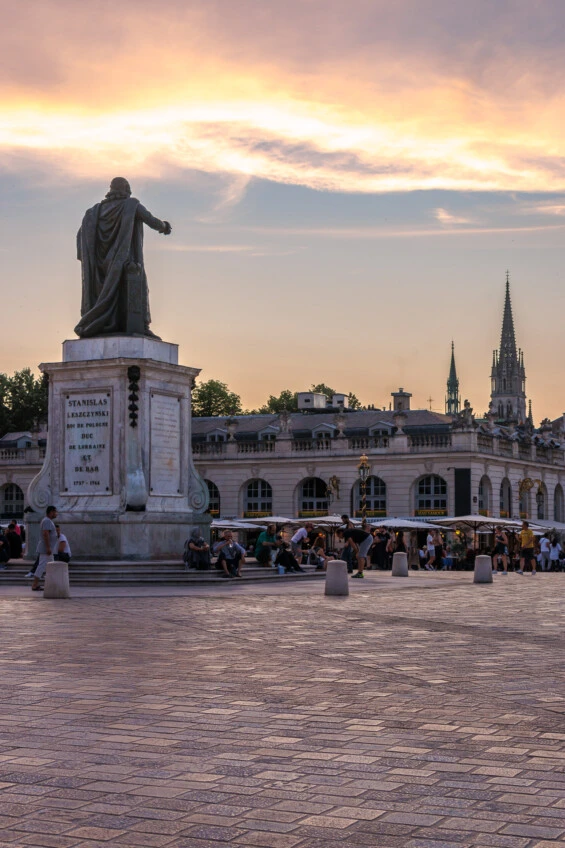 Cette photographie montre la statue de Stanislas Leszczynski, située sur la place Stanislas à Nancy, à l'heure du coucher du soleil. Le dos de la statue est tourné vers l'observateur, et le roi semble pointer vers l'horizon. Le ciel est teinté de couleurs chaudes et dorées, créant une ambiance douce et sereine.