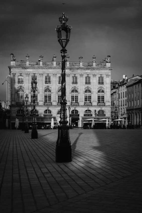 Cette photographie en noir et blanc montre une place pavée avec des lampadaires en fer forgé au premier plan. C'est la place Stanislas à Nancy.