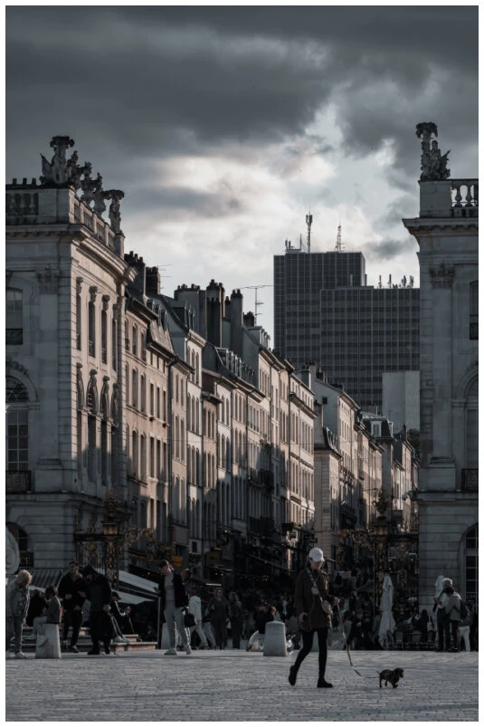 Cette photographie en couleur de la place Stanislas, montre une rue animée bordée de bâtiments historiques à l'architecture classique. Au premier plan, une personne marche avec un petit chien en laisse, ajoutant une touche de vie et de dynamisme à la scène.